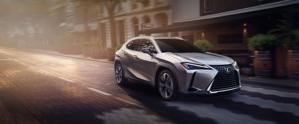 An all-new Silver 2024 Lexus UX driving through a downtown street. - Lexus of Huntsville, AL